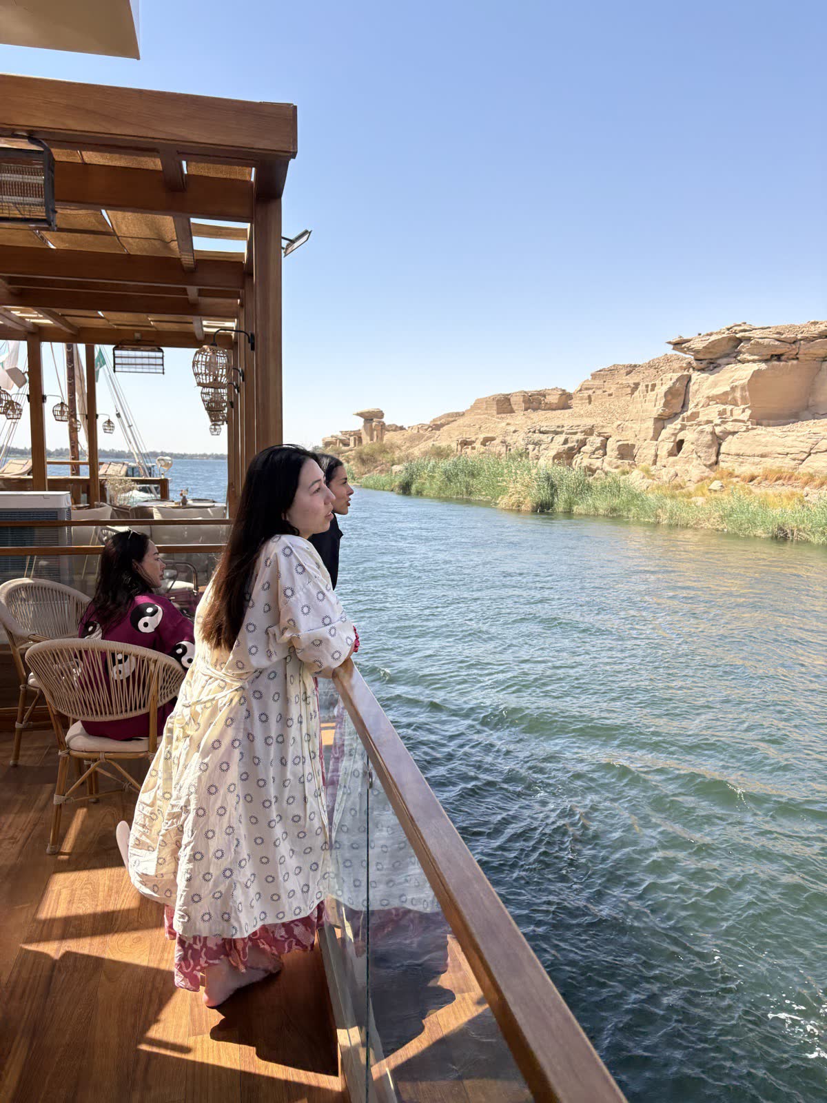A woman in a long floral dress looks out over the Nile from the deck of the dahabiya