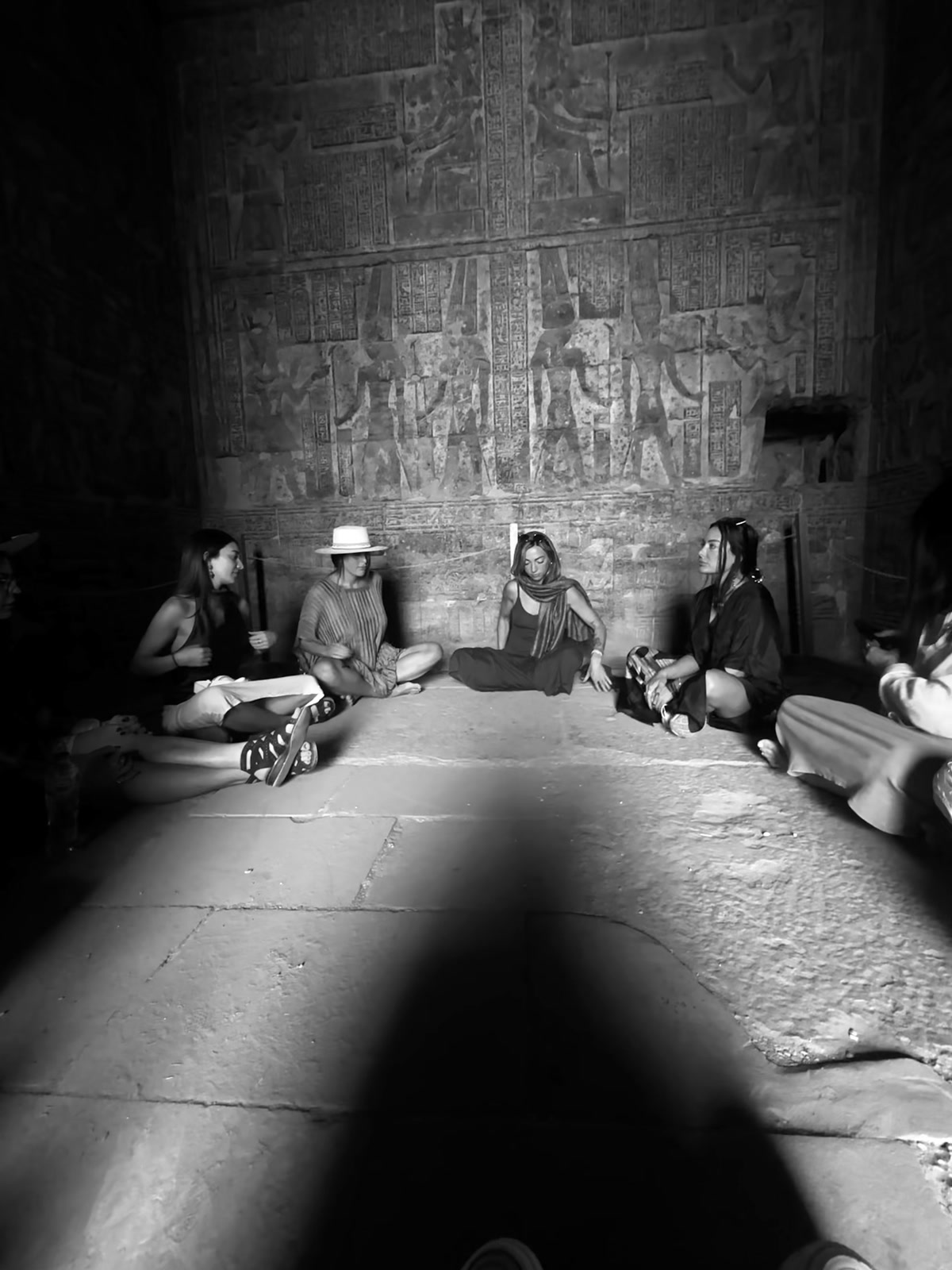 Women sit in a circle on the stone floor of a temple chamber, its walls carved floor to ceiling, in a single shaft of light